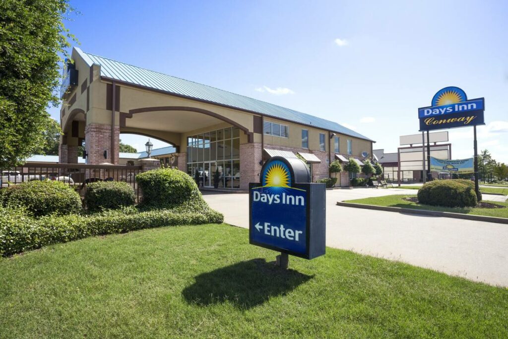 Days Inn in Conway, Arkansas has blue signs with a yellow sun above the letters. The motel is light red brick with a green roof and an entrance made of windows.