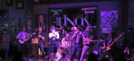 A band stands on stage at Kings in Conway, Arkansas. They are lit in purple light and a man and woman hold mics and sing.