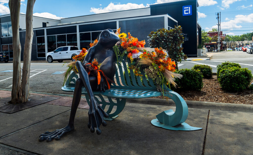 A large statue of a frog sitting on a blue iron bench in downtown Conway, Arkansas. He is looking sideways and has a corsage of bright autumn leaves and red ribbon tied around his wrist. There is a garland of red and yellow leaves and sunflowers draped along the back of the bench.