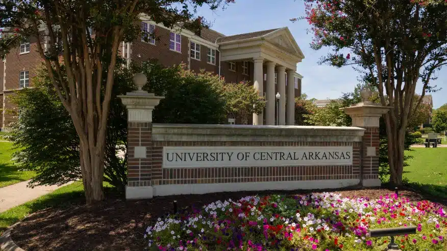 A brick sign saying 'University of Central Arkansas' in a flowerbed with bushes and purple flowers. A large brick classroom building with white columns is in the background in Conway, Arkansas.