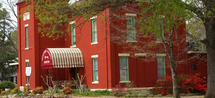 The Faulkner County Museum in Conway, Arkansas. The building is bright red and surrounded by flowerbeds with grass and red flowers. The covered entrance is white and red striped.