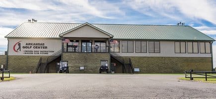 The front of the Arkansas Golf Center in Conway, Arkansas. A side paved driveway leads up to the building. Two metal staircases lead up the sides to enter the building with a green metal roof.
