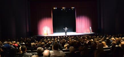 An announcer on state between two red curtains talking to an audience full of people at Reynolds Performance Hall in Conway, Arkansas.