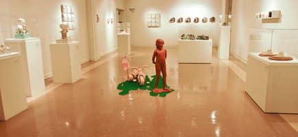 A display room in the Baum Gallery in Conway, Arkansas. The floor is shiny brown tile and the walls are a pale beige. There are white display cases lining the walls and a red statue of a girl with her pink tricycle in the middle.