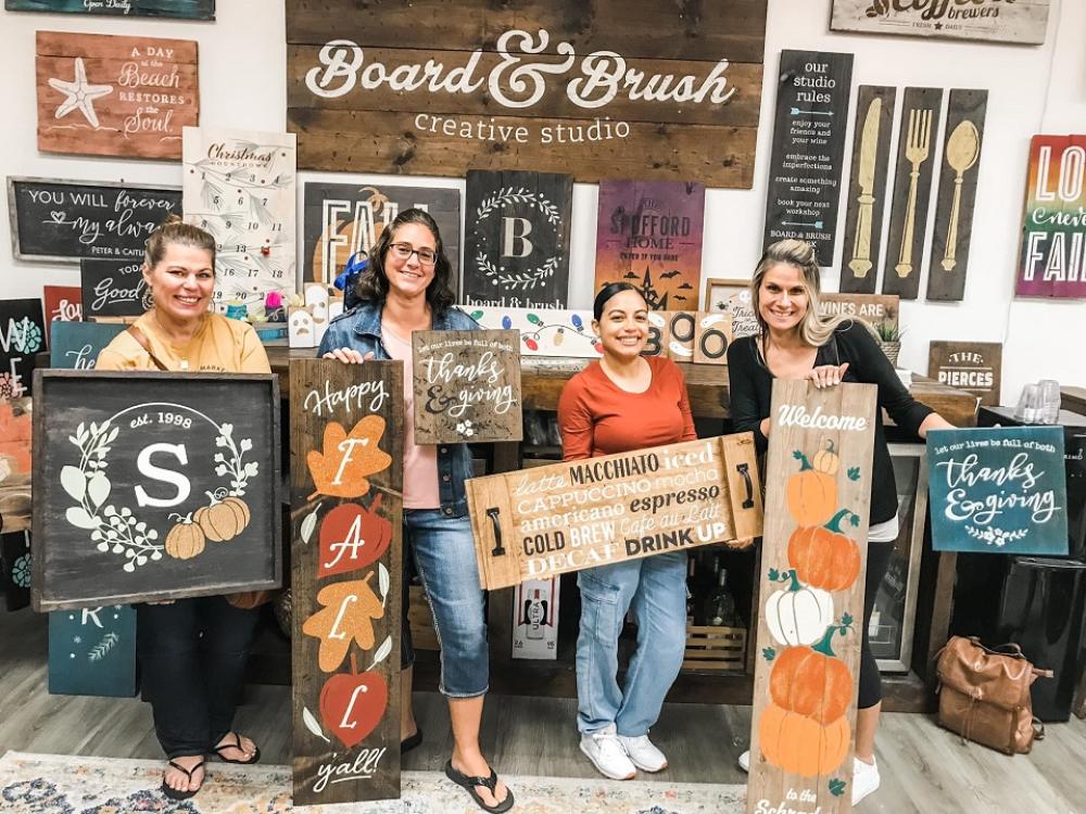 Four women pose in front of a wall of wooden signs and each is holding a wooden board that is Thanksgiving and fall themed. at Board & Brush Creative Studio in Conway, Arkansas.
