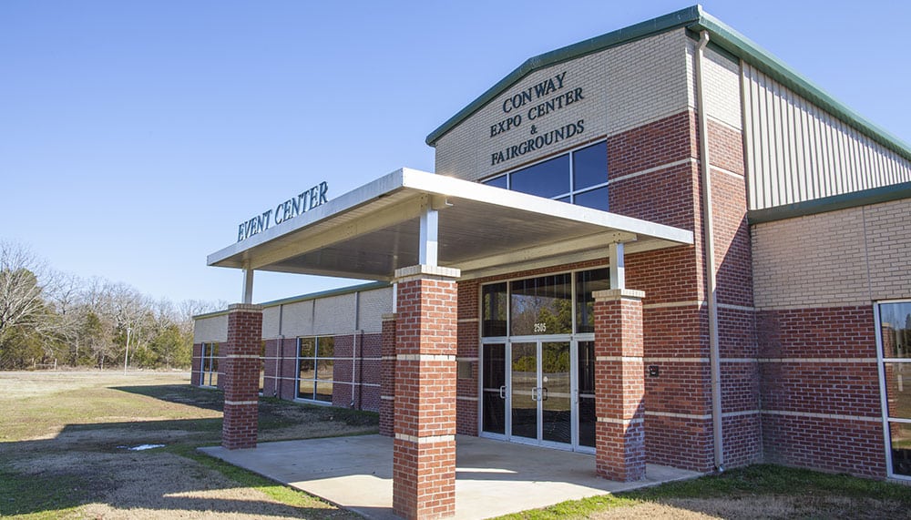 A building with a mix of red and white brick in Conway, Arkansas and the covered entrance has a sign above it that says "Conway Expo Center & Fairgrounds." There are four glass doors at the entrance.