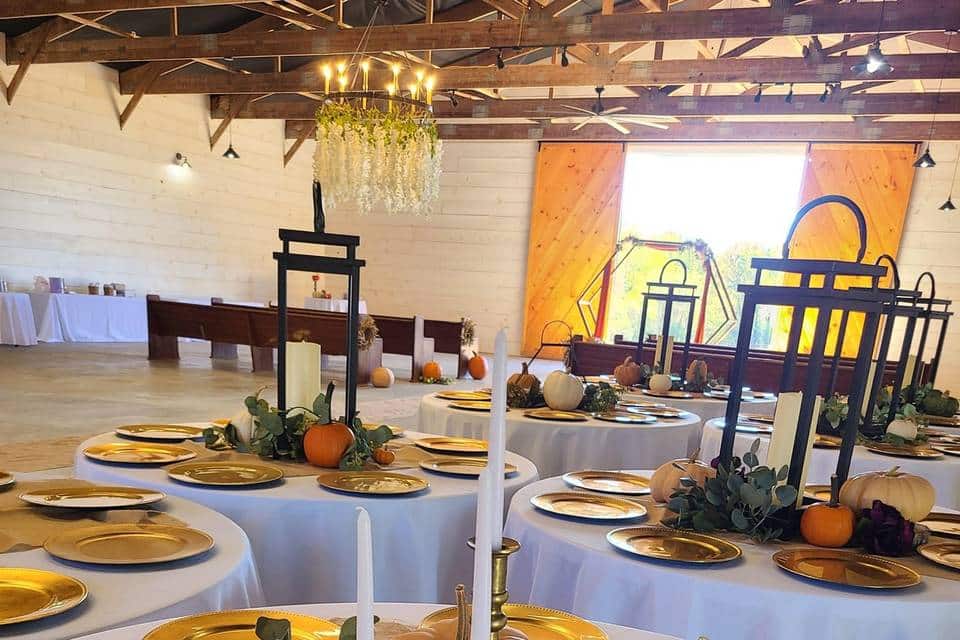 A white brick open room with big wooden barn doors at The Pines Event Center in Conway, Arkansas. The room is decorated for fall with circle tables covered in white tablecloths with candles and pumpkins.