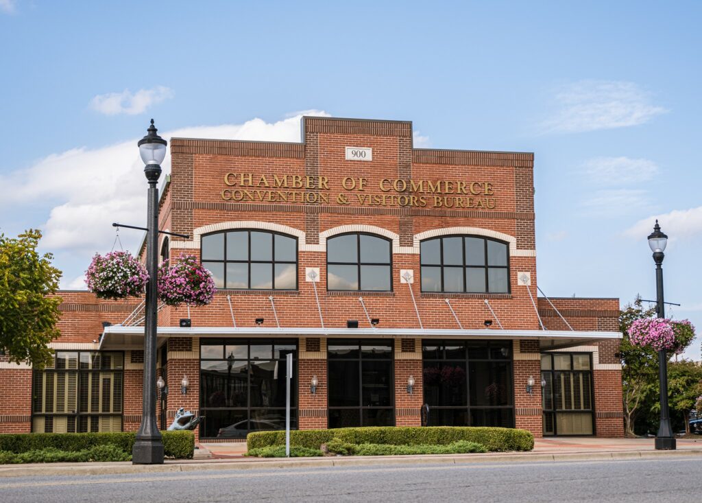 A red brick building with a gold sign that says "Chamber of Commerce Convention & Visitors Bureau" in Conway, Arkansas. There are two street lamps with pink baskets of flowers in front.