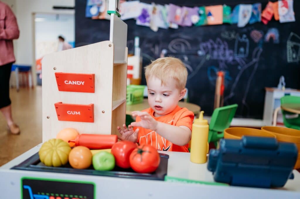A little blond boy in an orange Nike shirt reaches for a plastic bottle of ketchup that is laying on a toy conveyor belt with toy tomatoes and other fruit at the Share the Love Kids Club in Conway, Arkansas.