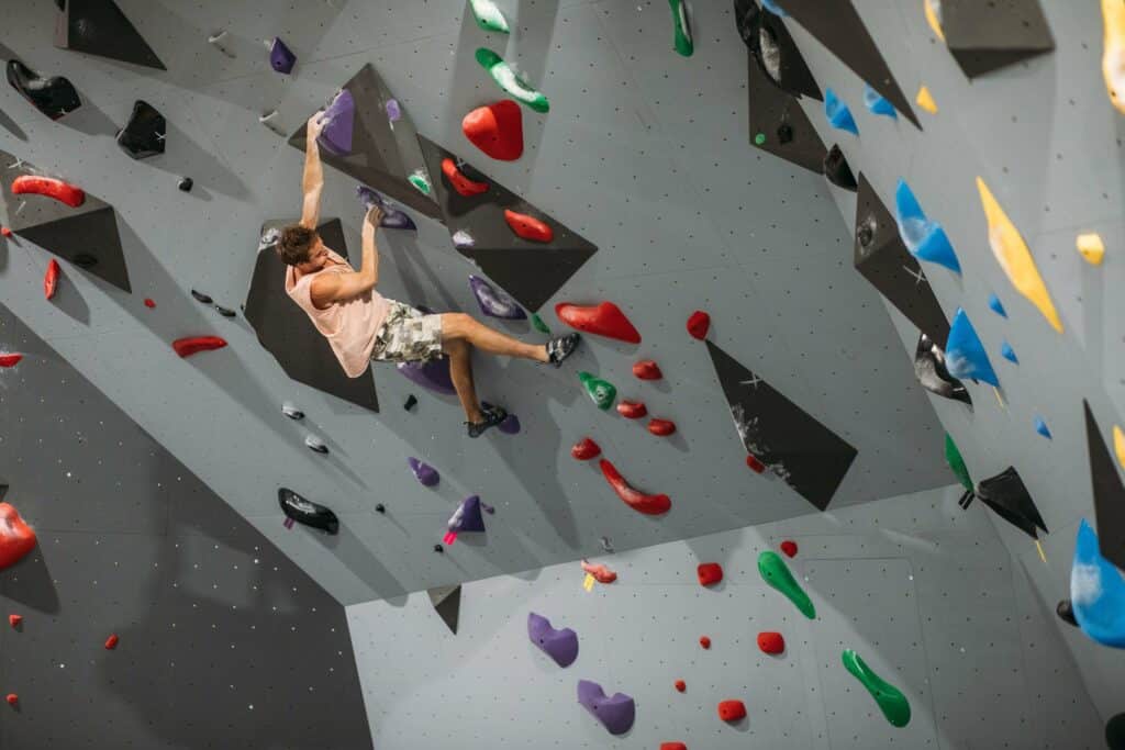 A man in a peach colored tank top and climbing shoes hangs from a purple hand hold. The gray climbing wall is scattered with red, blue, yellow, green, black, and purple holds at Climb Conway in Conway, Arkansas.