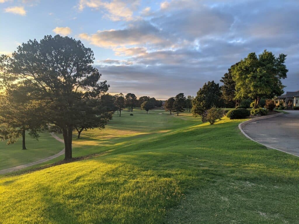 Alarge green golf course spotted with trees at the Conway Country Club in Conway, Arkansas. The sky is blue with many clouds.