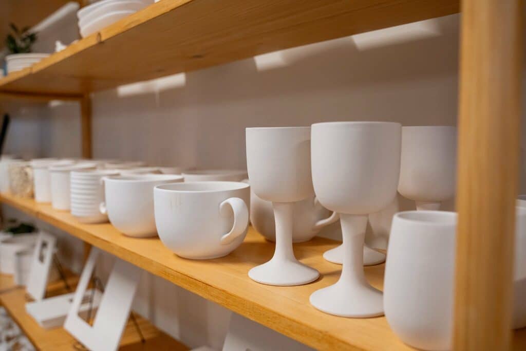 A wooden shelf holds mugs and goblets of unpainted white pottery at Glazin Pottery Studios in Conway, Arkansas.