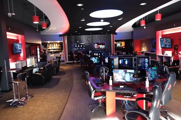 A dimly lit room has red gaming desks set up in a line in the center of the room with monitors and speakers. There is a snack bar and lounge chairs in front of a tv off to the side in Spec Ops Gaming Lounge in Conway, Arkansas.