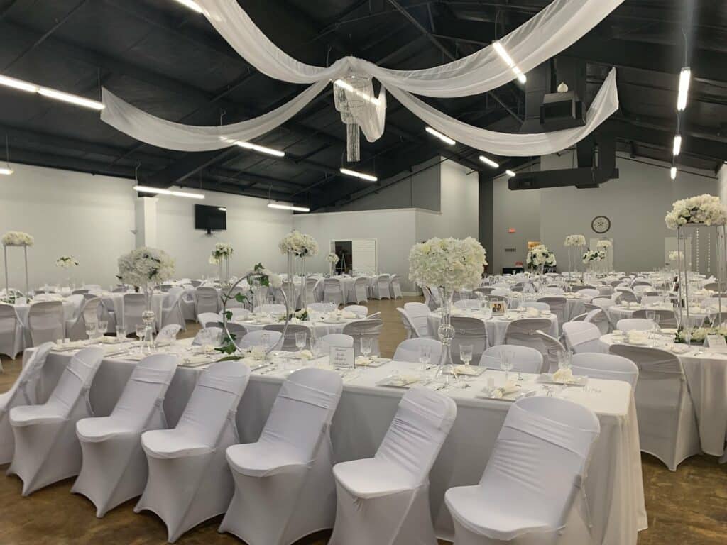 A large event room with rectangular and circular tables at the Willow Event Center in Conway, Arkansas. Tables are set with tall flower vases and glass place settings. Chairs and tables are covered in white cloth.