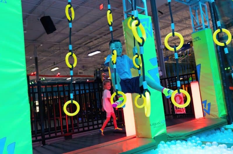 A young boy climbs on neon yellow rings. He is dangling above a ball pit of light blue plastic balls at Urban Air Trampoline & Adventure Park in Conway, Arkansas.