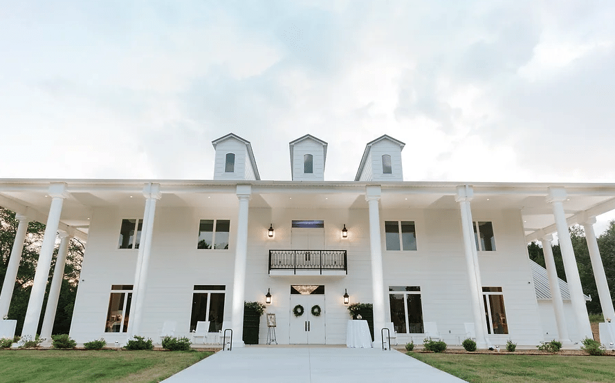 An impressive large white building has six tall columns and a double door entrance with two wreaths. There is a small iron balcony hanging above the front doors. The building is Legacy Acres in Conway, Arkansas.
