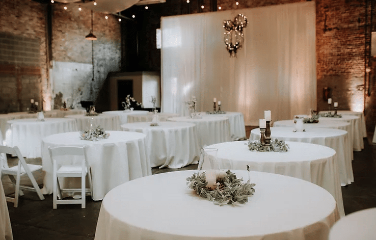 A room with red brick walls and a hanging white curtain at The Brick Room in Conway, Arkansas. There are several circular tables with white chairs and white tablecloths. There are a few candles and some greenery in the center of each table.